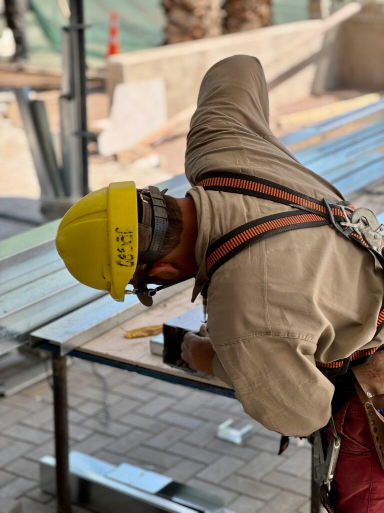 A construction worker in safety gear measuring a metal beam outdoors on a sunny day.